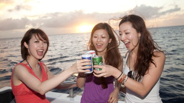 Three women drinking beer and cheering to the amazing time they are having as the sun sets behind them over the water in Tumon, Guam