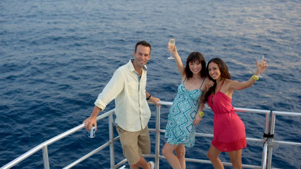 A man and two women drinking on the Big Sunset Cruise as the sun sets over the blue water in Tumon, Guam