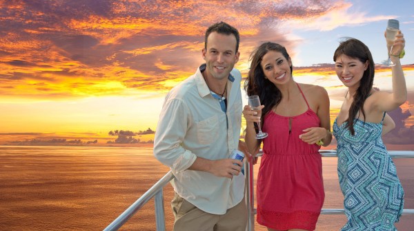 A man with a beer and two women with drink at the edge of the deck on the Big Sunset Cruise with the sunset in the background