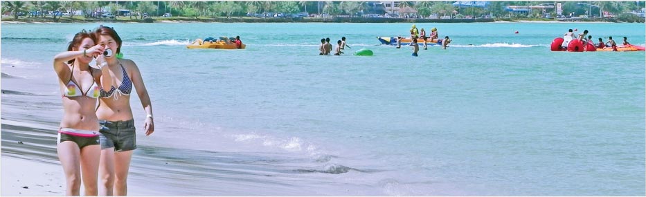 safety-banner Two women walking along the beach in Guam taking photos while people enjoy water activities in the background