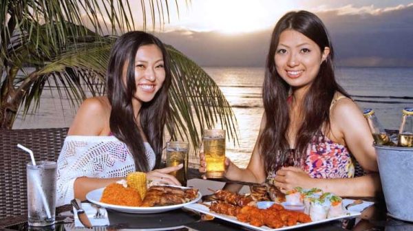 Two women enjoying BBQ and beer beachside while the sun sets over the ocean in the background in Tumon, Guam