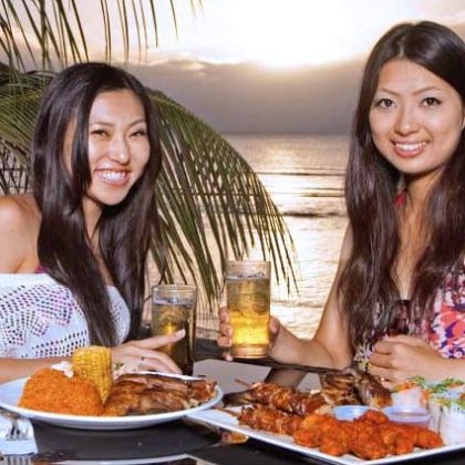 Two women enjoying BBQ and beer beachside while the sun sets over the ocean in the background in Tumon, Guam