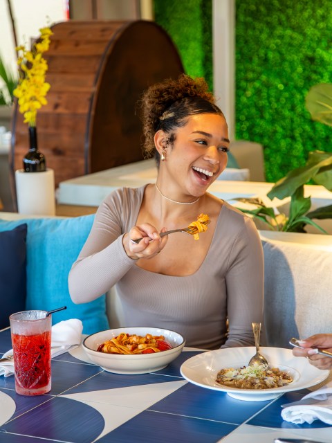 Smiling woman eating pasta at a restaurant, with drinks and a salad on the table.