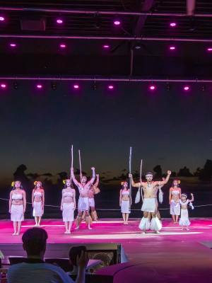 Performers in traditional attire with props on pink-lit stage at sunset.