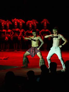 Two male dancers perform on stage with a group of dancers in red headdresses behind them.