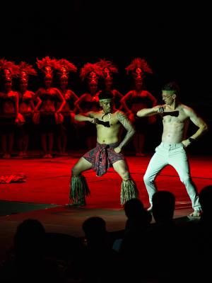 Two male dancers perform on stage with a group of dancers in red headdresses behind them.