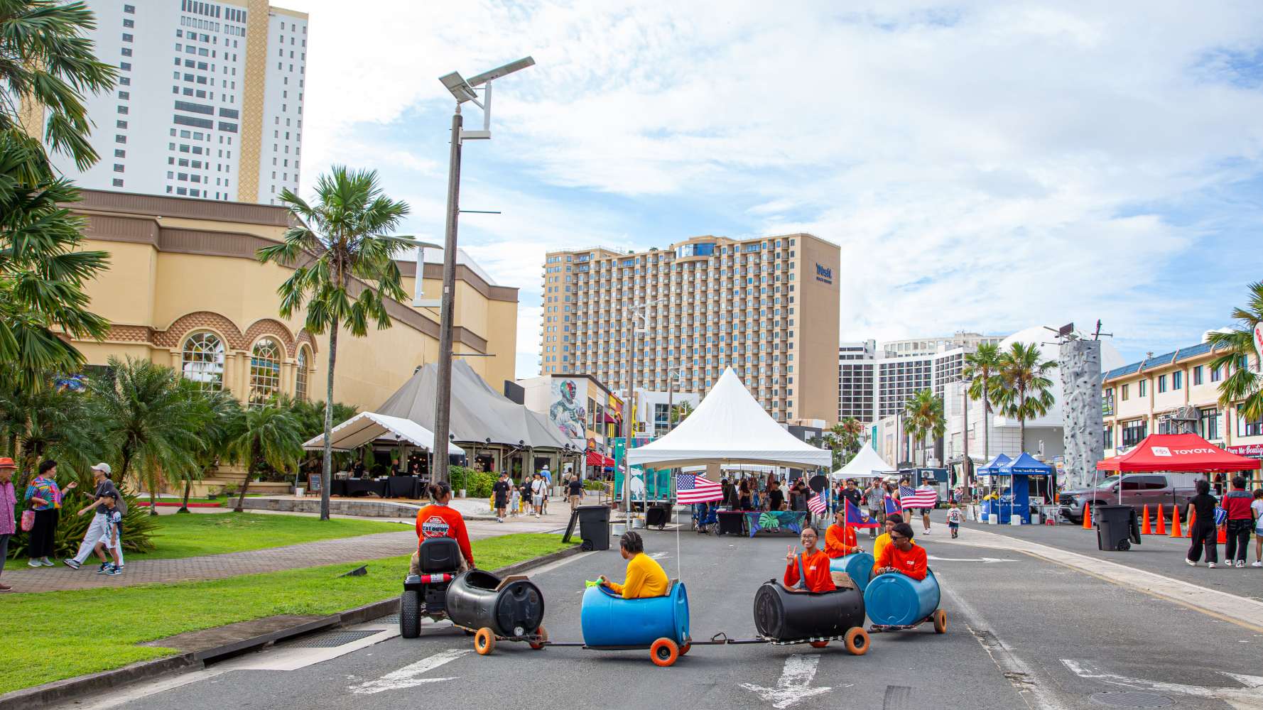 Urban festival with people on barrel train, tents, and high-rise buildings in the background.