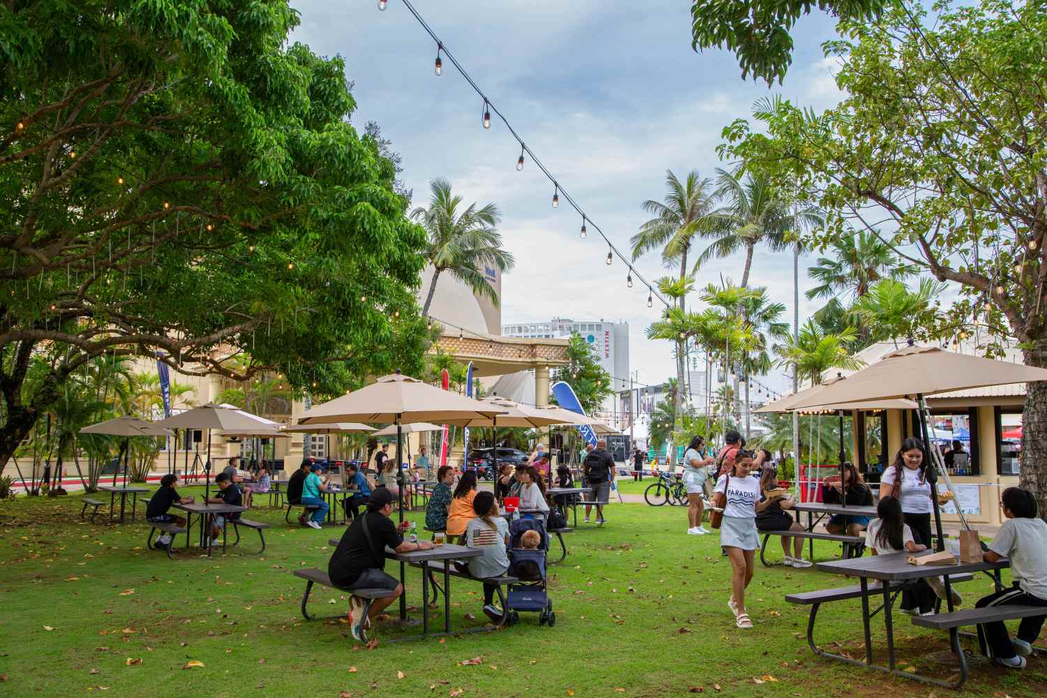 People sitting at picnic tables under trees and umbrellas in a park setting.