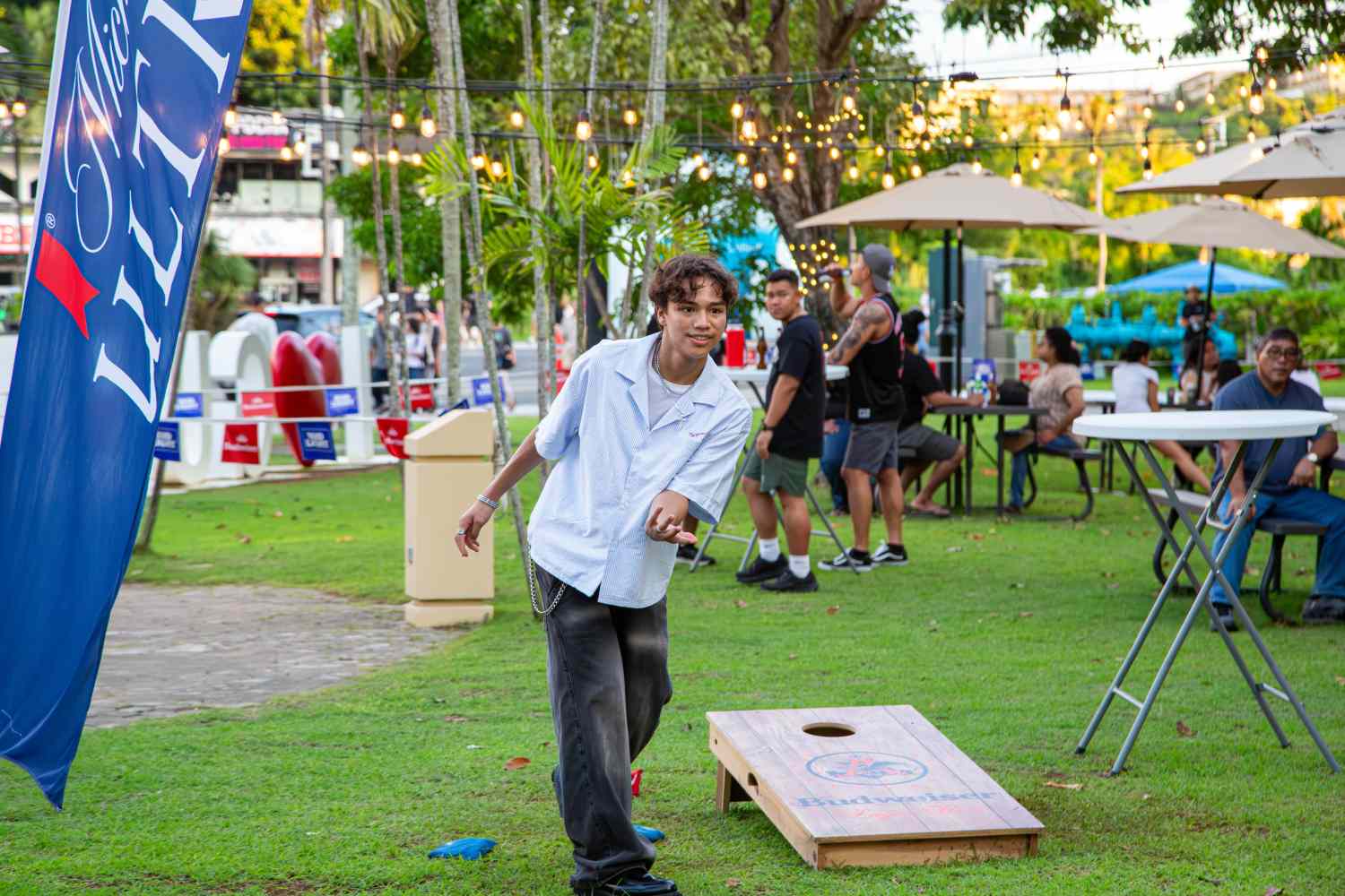 Person playing cornhole outdoors at an event with tables and string lights.