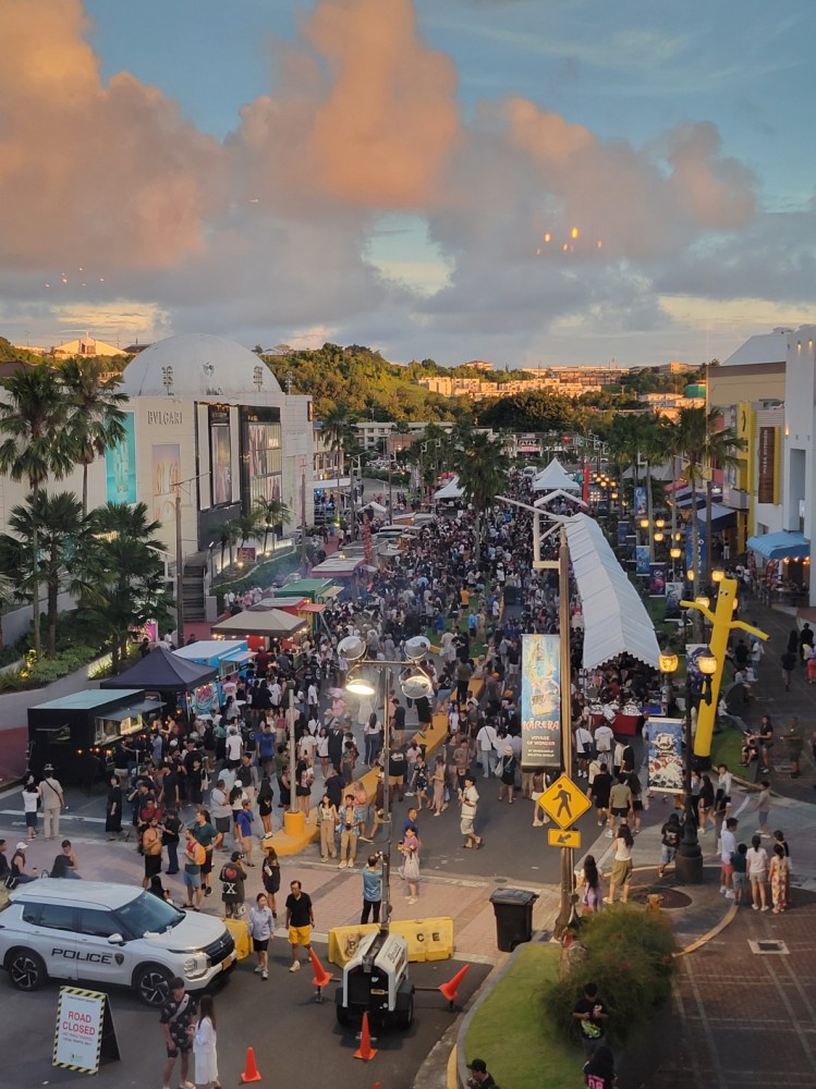 Large crowd at outdoor event with street vendors and police vehicle under a cloudy sky.