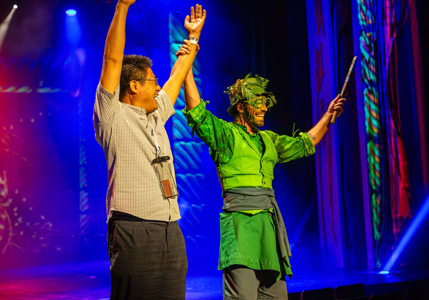 Two people on stage celebrating, one in green costume with leafy accessories, colorful lighting background.