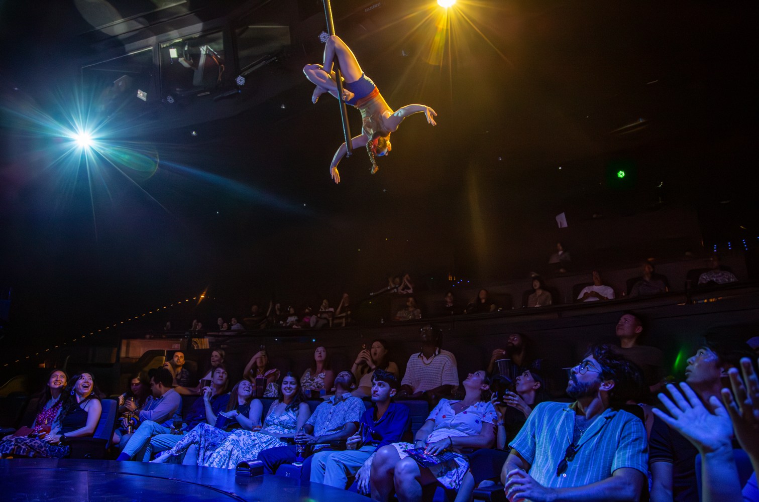 Aerial performer on a pole above an audience in a dimly lit theater.