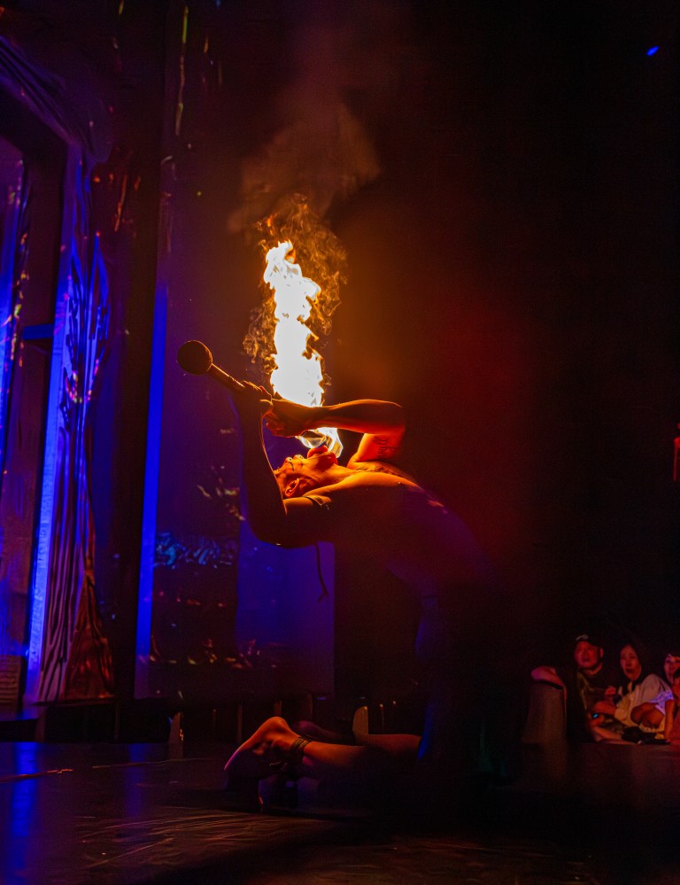 Performer kneeling and breathing fire on stage in dark, colorful setting.