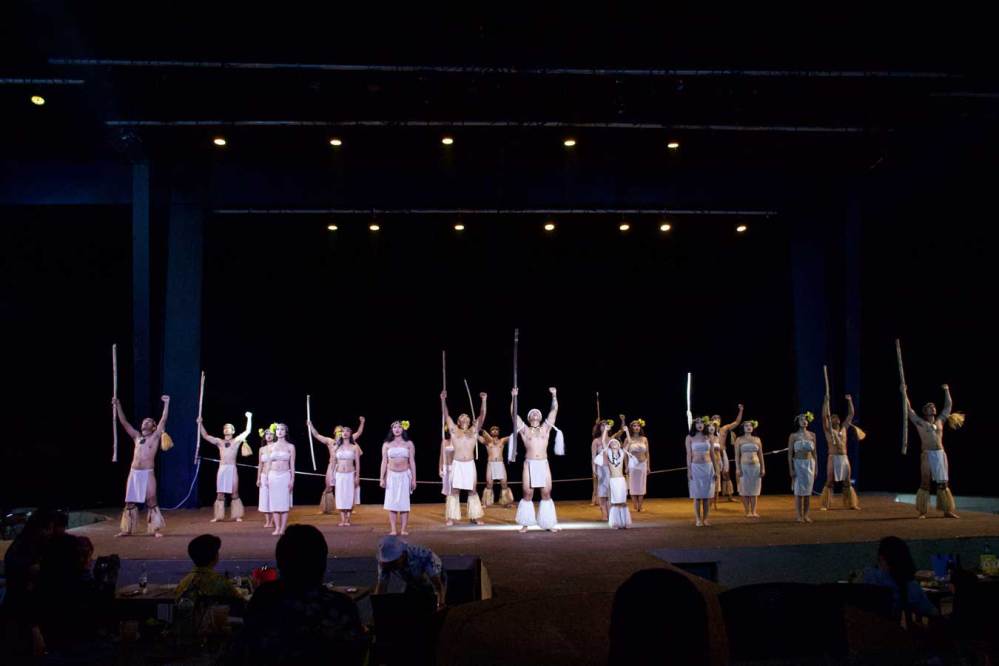 Group of performers in tribal costumes with staffs on stage under spotlights.
