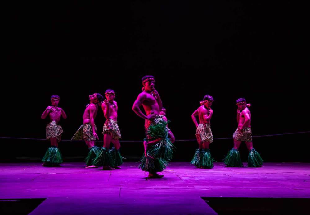 Six performers in grass skirts dance on stage under purple lighting.