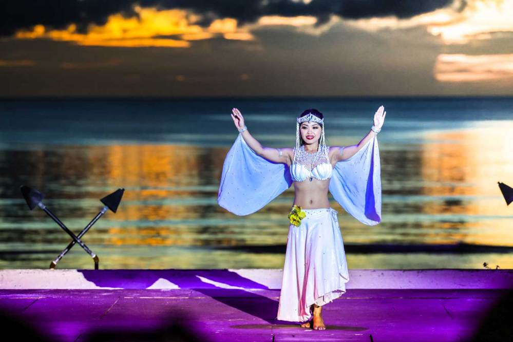 Dancer in white outfit performing on stage by the ocean at sunset.