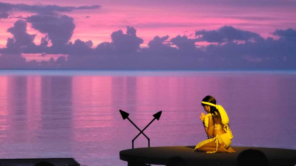 Person in yellow costume kneeling on stage with purple sunset over water.