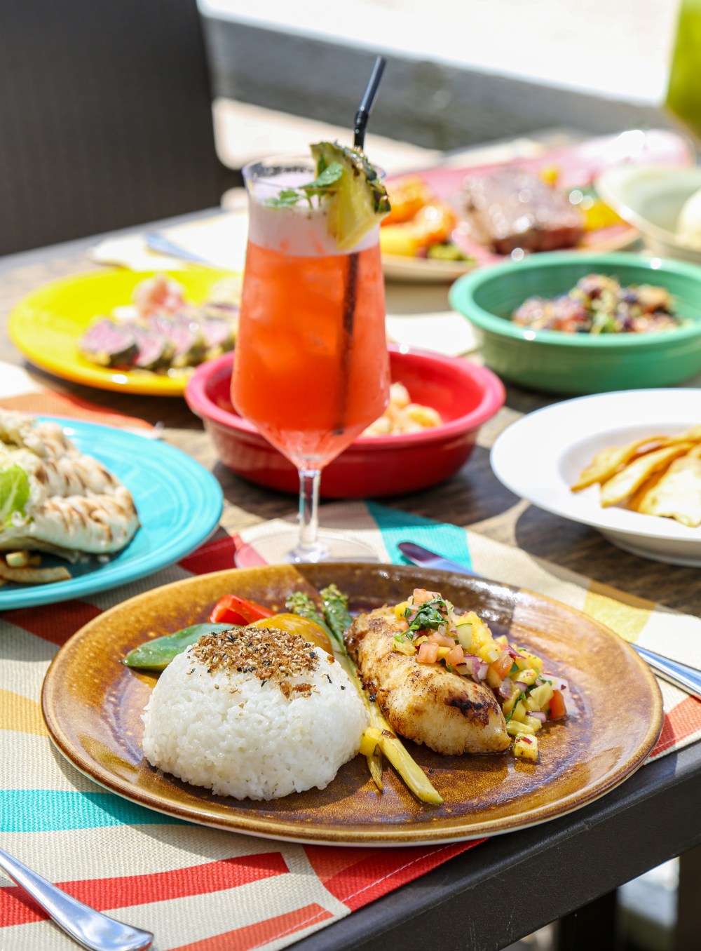 Plate with rice and fish next to a tropical drink and colorful dishes on a table.