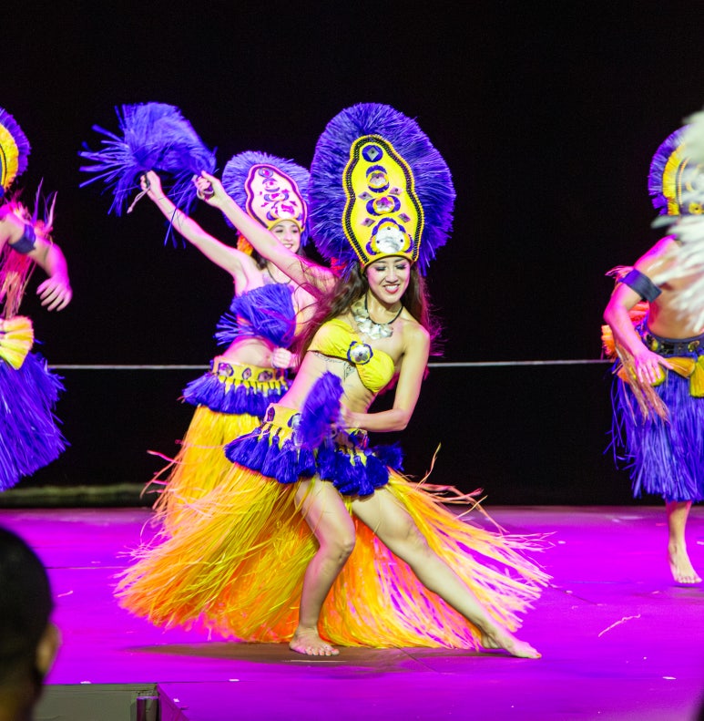 Dancers in bright costumes perform on stage with colorful headdresses and skirts.