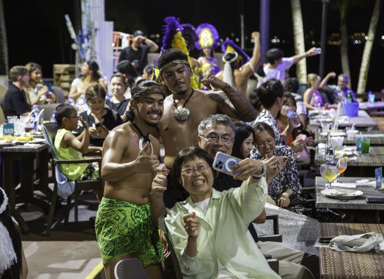 People taking a group selfie at an outdoor event with dancers in the background.