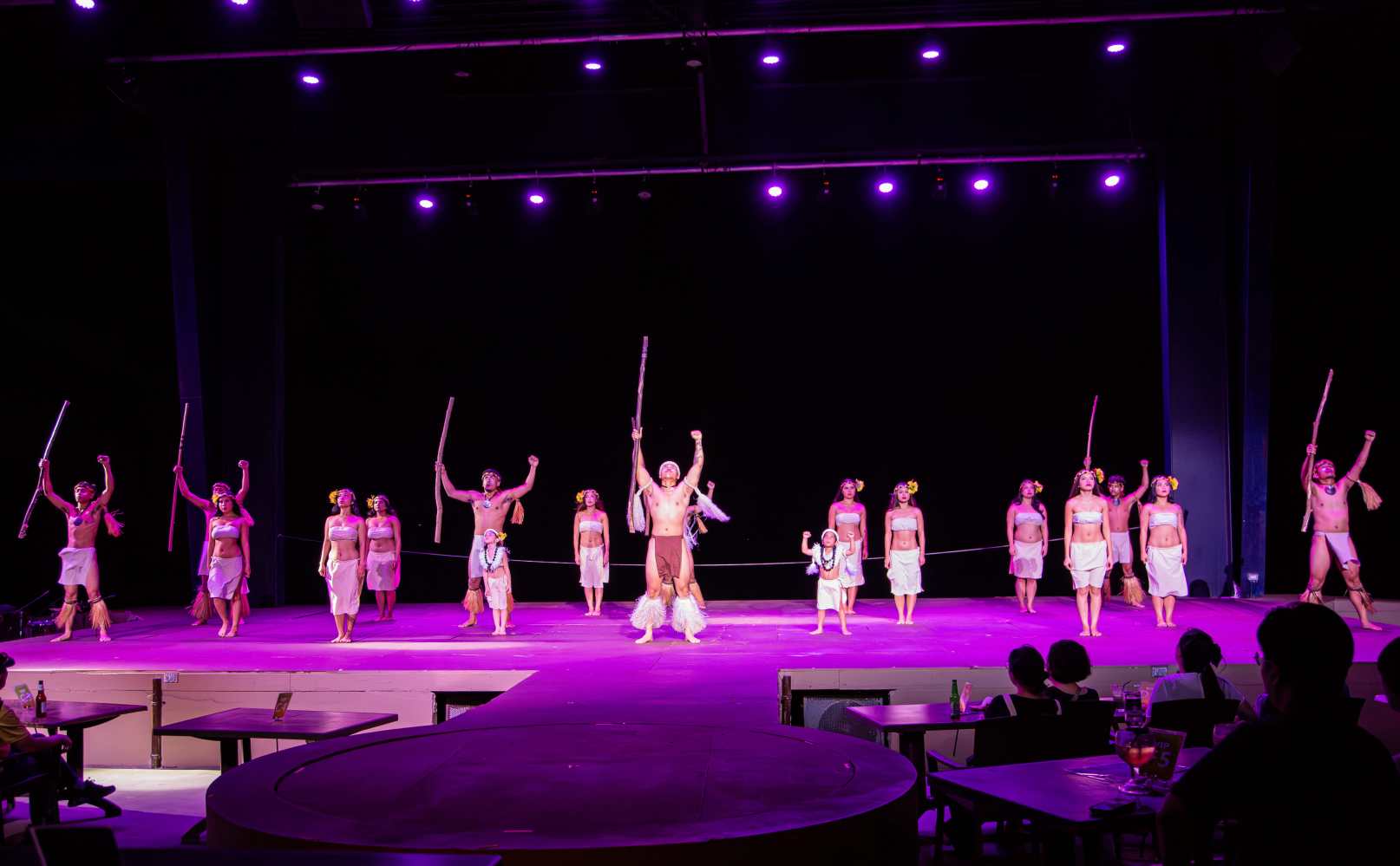 Performers in tribal costumes dance with sticks on a stage under purple lighting.