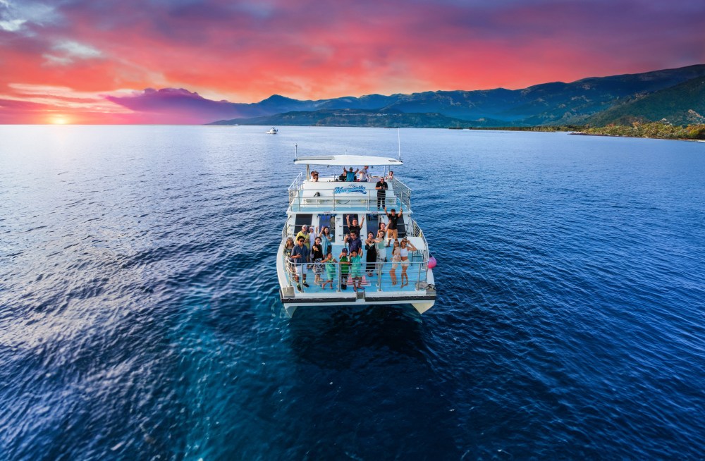 A boat full of people on a blue ocean with a vibrant sunset sky and distant mountains.