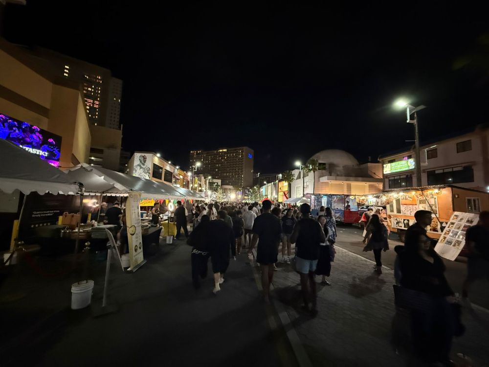 Night market with food stalls and people walking on a city street.
