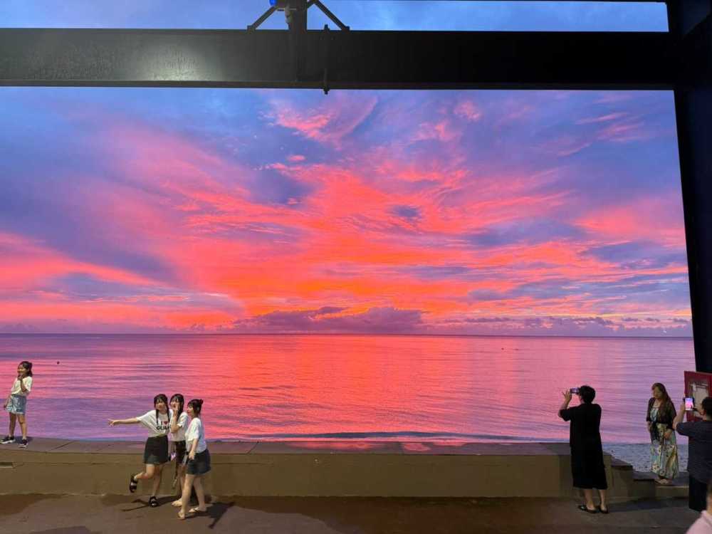 People photographing a vibrant pink and orange sunset over a calm ocean, framed by a dark structure.