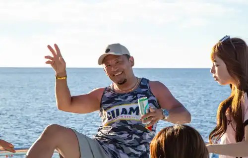 Smiling man in tank top and cap holds a drink, posing on a boat with an ocean backdrop.
