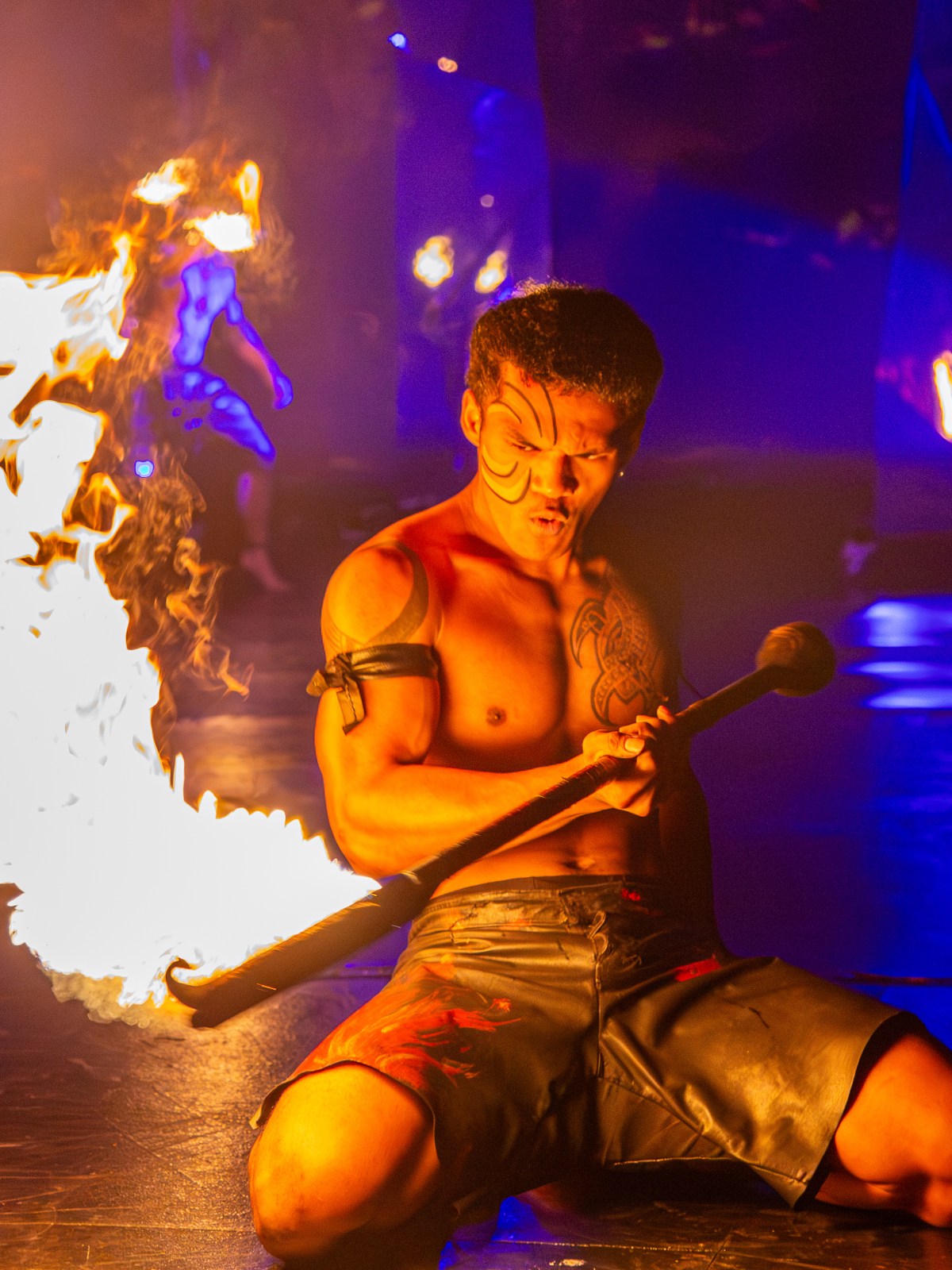 A shirtless performer kneels with a burning torch against a colorful, dimly lit backdrop.