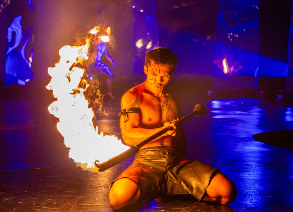A shirtless performer kneels with a burning torch against a colorful, dimly lit backdrop.