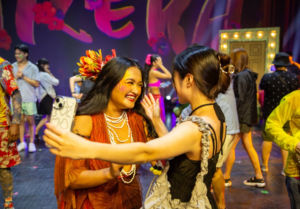 Two women smiling and taking a selfie at a colorful, lively event with people in the background.