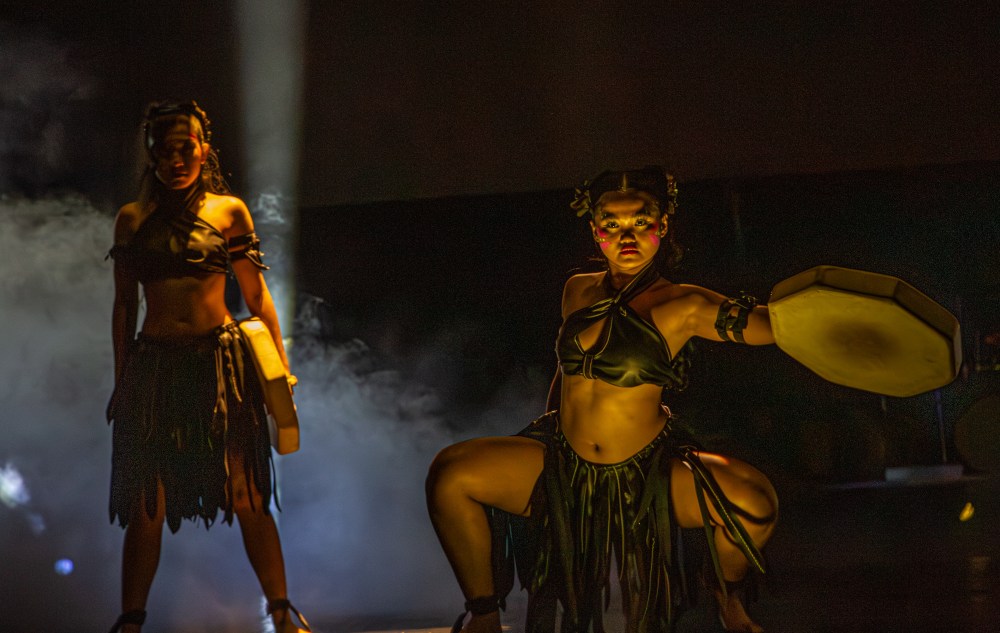 Two dancers in tribal attire perform on stage, holding drums under dramatic lighting.