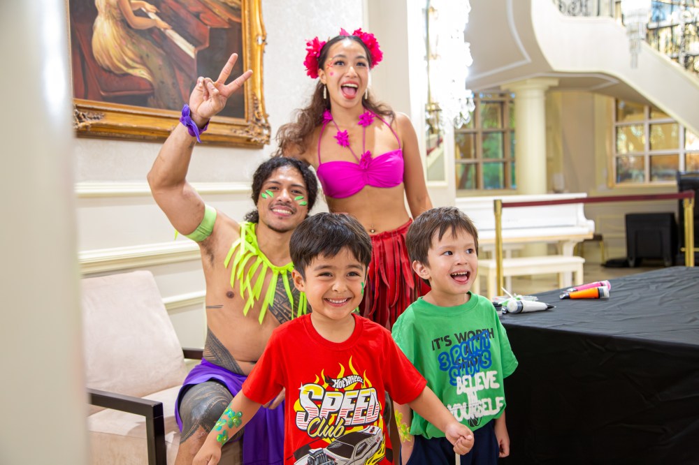 Two children smiling with performers in colorful costumes in a bright indoor setting.