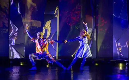 Two dancers pose dramatically on stage with colorful lighting and mirrored backgrounds.