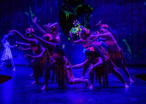 Group of dancers in vibrant costumes performing on a dimly lit stage with blue lighting.