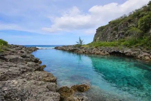 Rocky lagoon with clear blue water and green shrubs under a partly cloudy sky.