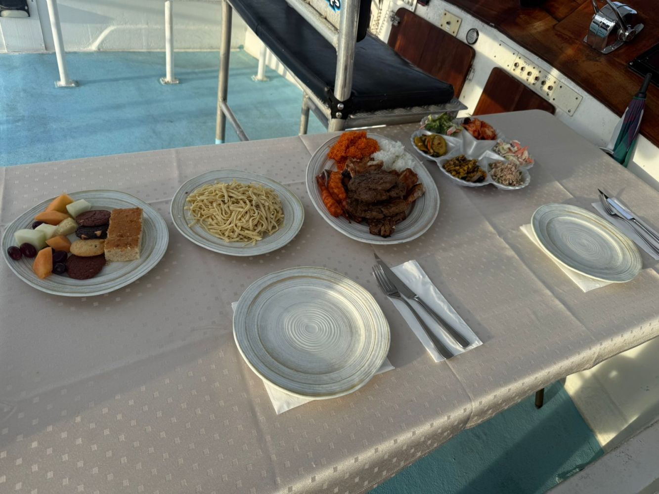 Table with assorted dishes including noodles, fruit, meat, and side dishes on a beige tablecloth.