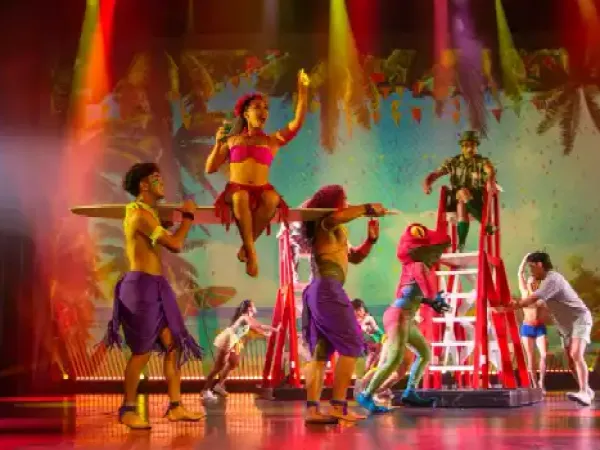Performers in colorful outfits on stage with surfboards and tropical backdrop.