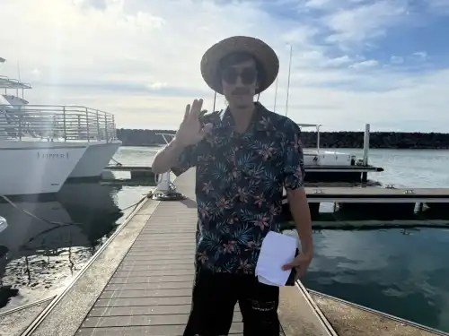 Person in floral shirt and straw hat waves on a dock by boats under a cloudy sky.