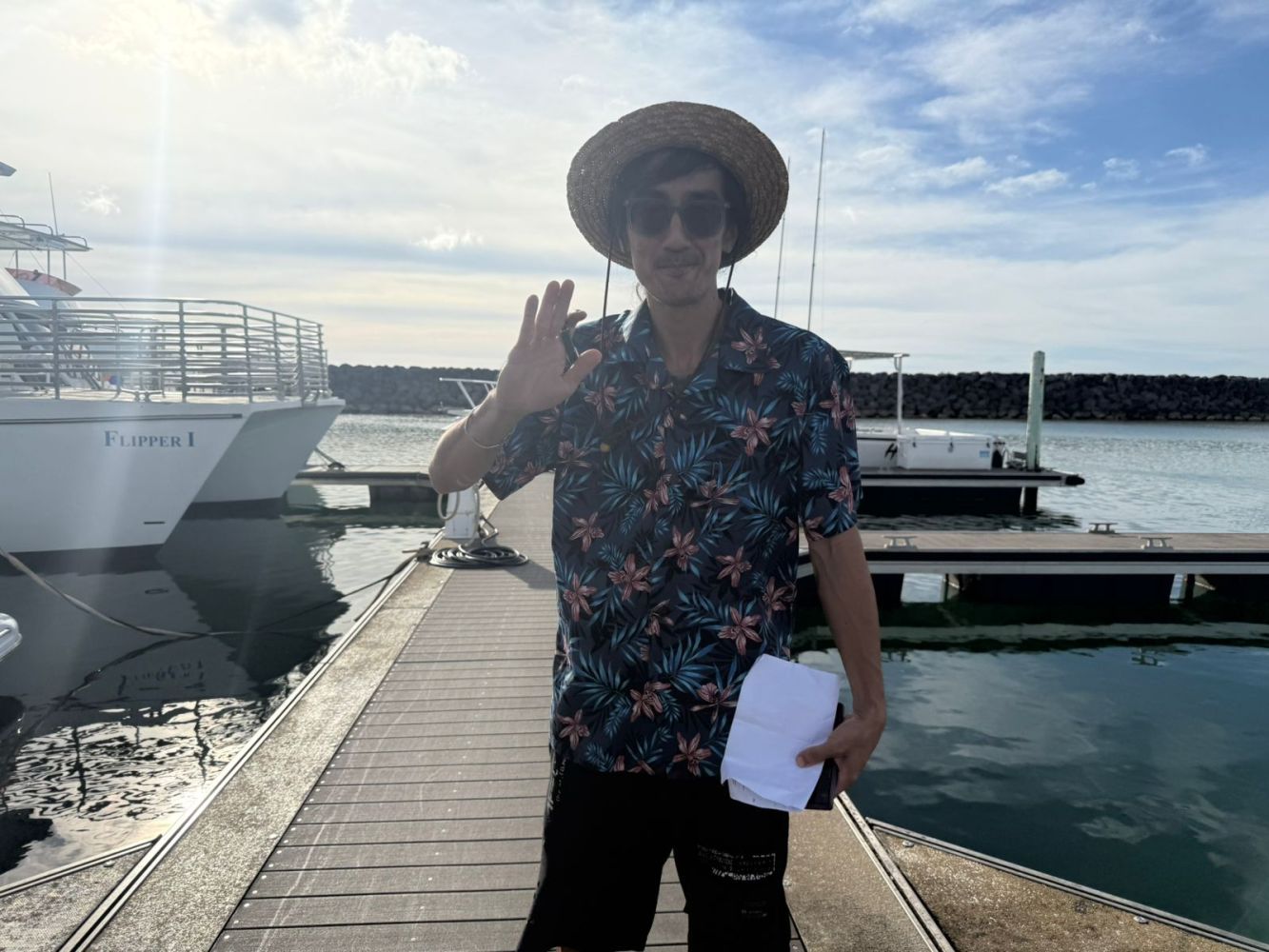 Person in a floral shirt and straw hat waving on a dock near boats.