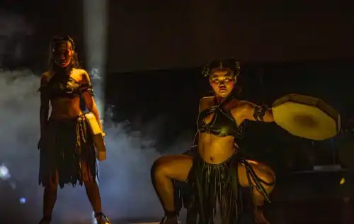 Two performers in tribal attire on stage with dramatic lighting and smoke.