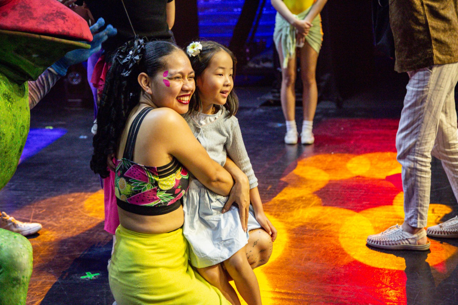 Woman hugging a child on stage, colorful lighting and people in background.