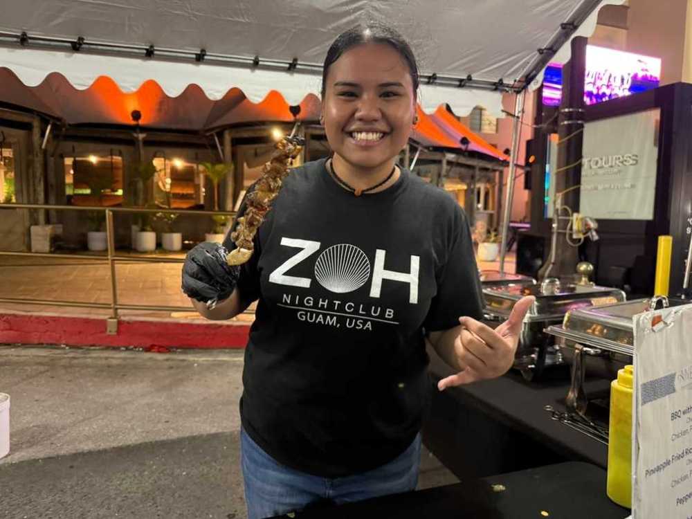 Smiling person holding a skewer at a food stall, wearing a ZOH Nightclub shirt.