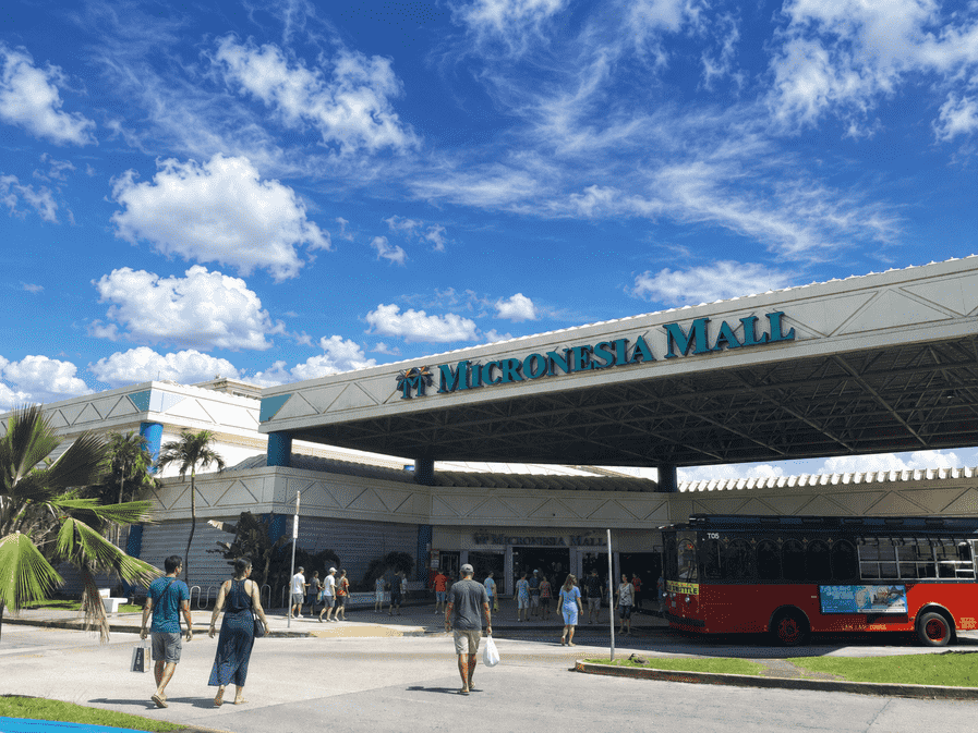 People entering Micronesia Mall with a red bus parked outside under a partly cloudy sky.