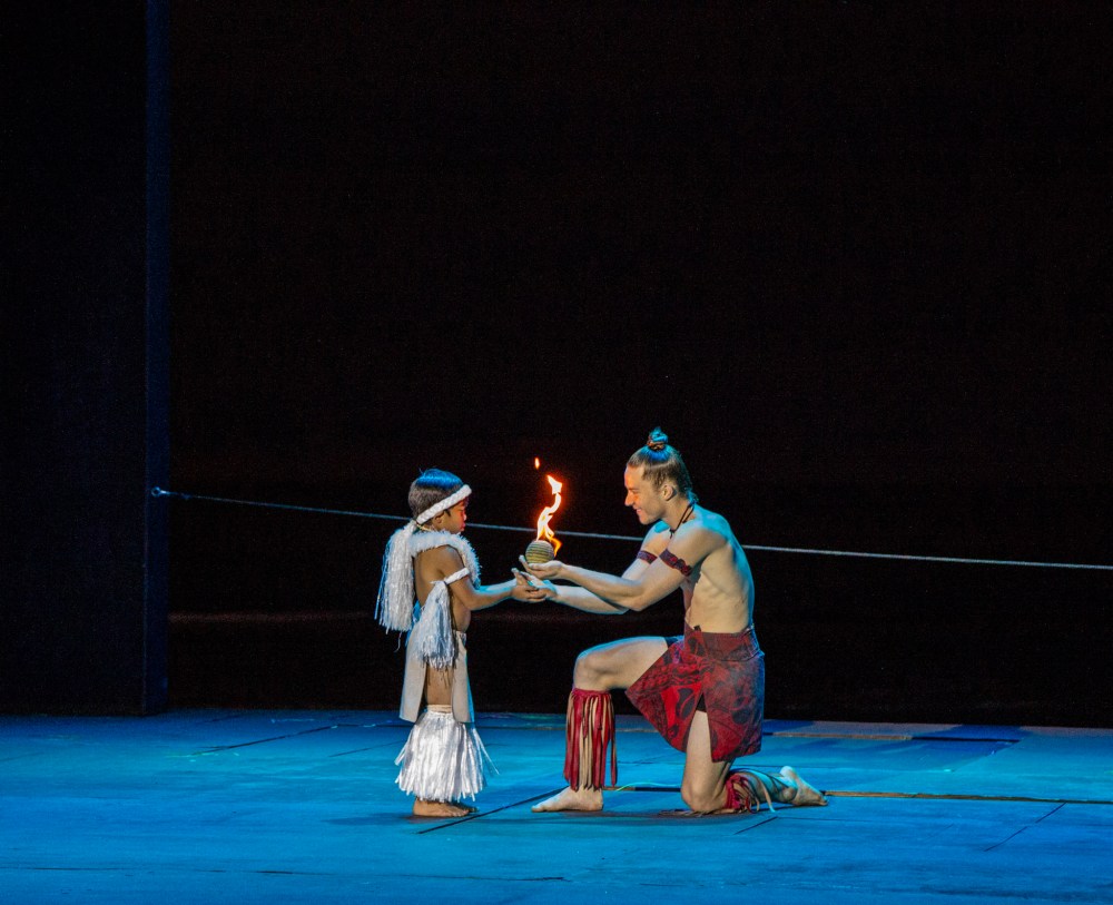 Two performers on stage; one kneels, holding a flame, wearing tribal attire.