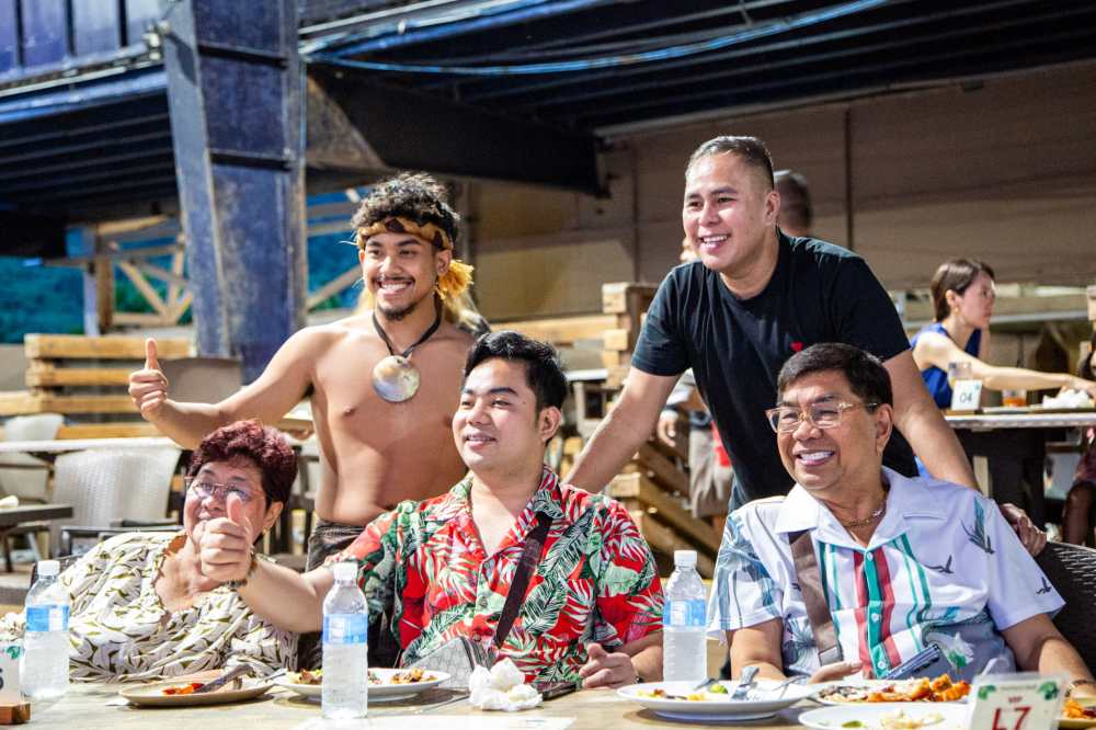 Group of five people smiling at an outdoor table, one shirtless in a traditional necklace.