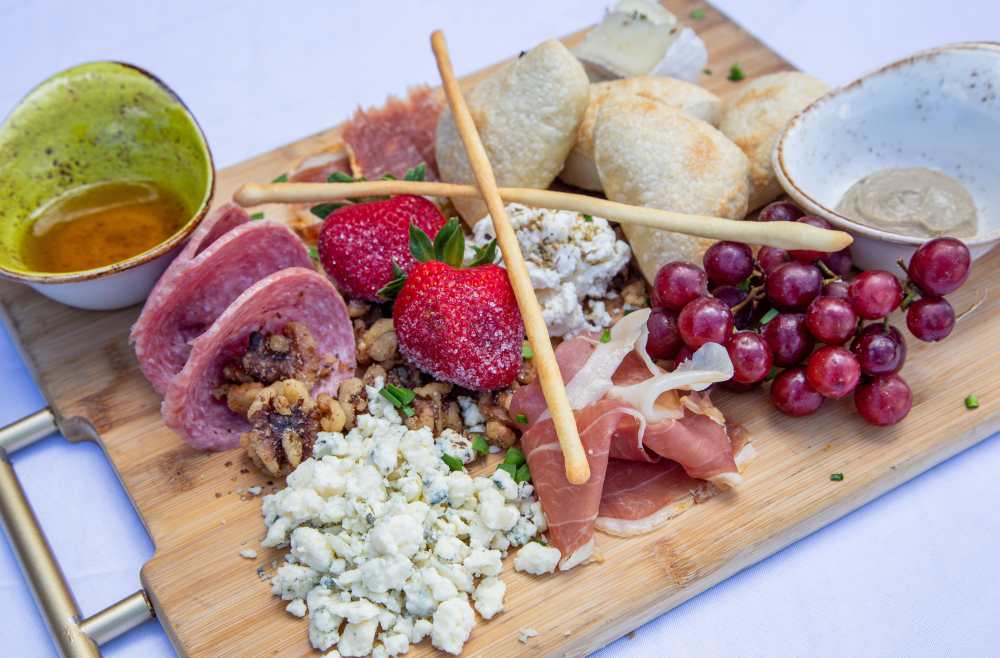 우드 도마 플래터 위에 풍성하게 차려진 지중해식 샤큐테리(Charcuterie)와 치즈 플래터. 얇게 썬 프로슈토와 살라미, 블루치즈, 신선한 딸기와 포도, 그리시니 과자 스틱, 따뜻한 식전 빵과 소스들이 다채롭게 올려져 있어 와인이나 칵테일 안주로 완벽한 구성을 보여준다.