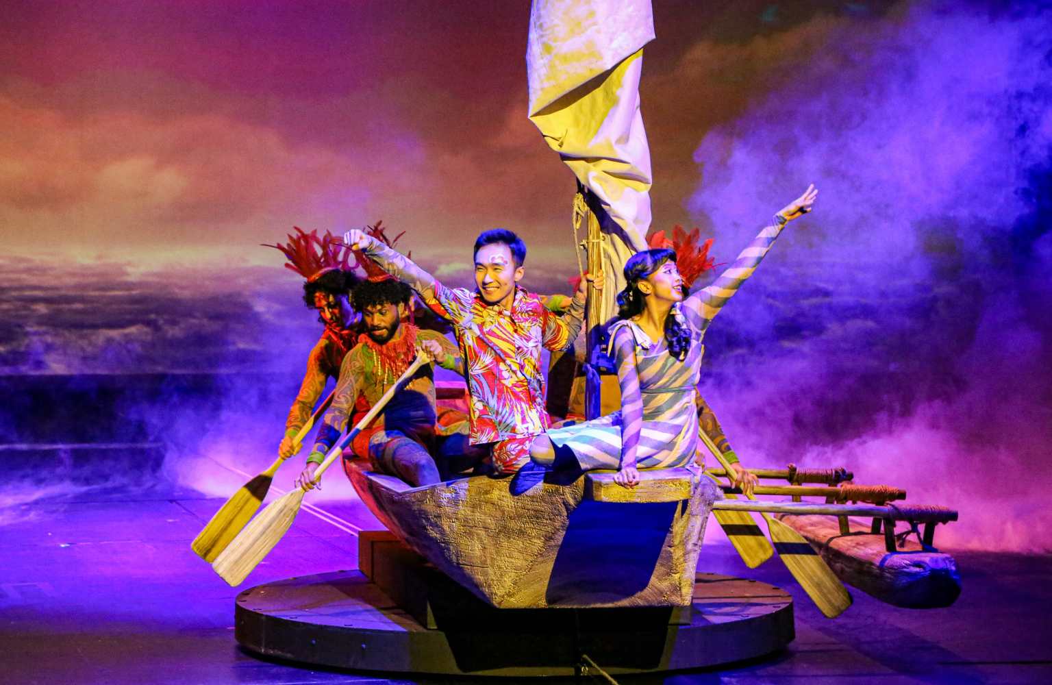 Three performers in a boat on stage with a dramatic background.