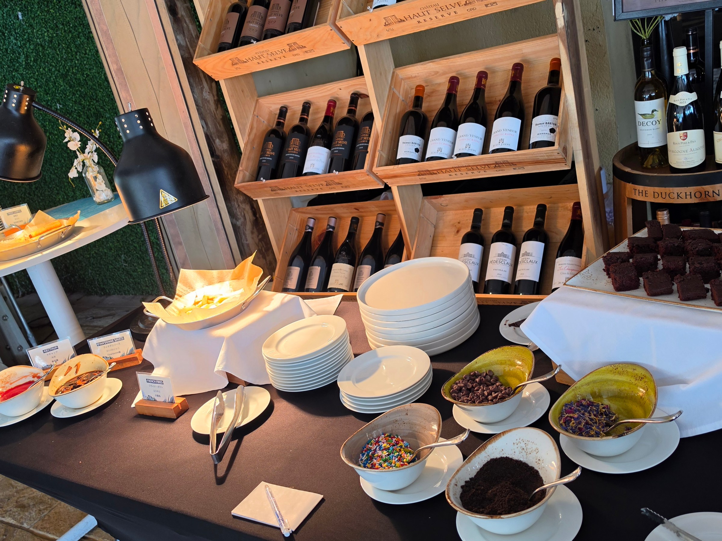 Buffet setup with wine, plates, desserts, and toppings in a cozy setting.